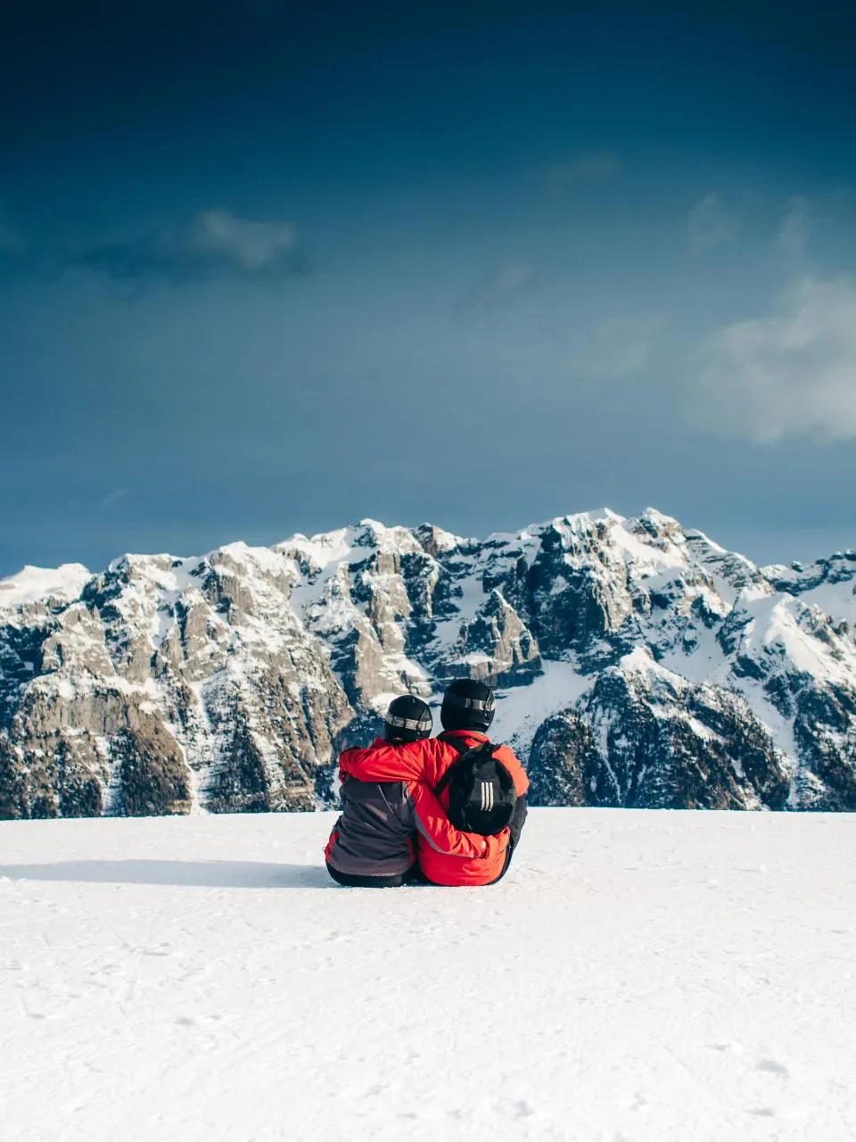 Pareja con casco de esquí sentada en la nieve frente a las montañas